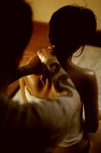 Zdjęcie przedstawia Tomka Raszewskiego - masażystę z wieloletnim doświadczeniem oraz właściciela Mahasanti.pl, który prowadzi sesję ajurwedyjskiego masażu jogicznego i masuje lewe ramie siedzącej klientki.